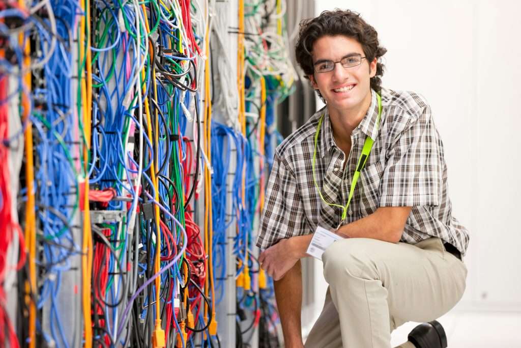A young man with glasses is kneeling beside a wall of tangled network cables in a server room. He is smiling and wearing a name badge and a lanyard.
