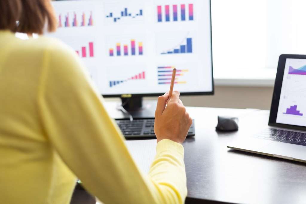A person wearing a yellow sweater is seated at a desk, holding a pencil and pointing towards a computer screen displaying various bar graphs and charts, suggesting data analysis or statistics work.