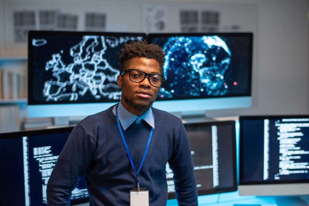 A person stands in front of multiple computer monitors displaying coding and data visualizations. The setting appears to be a tech-focused workspace.