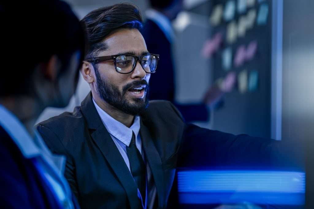 A man in a suit is engaged in a discussion while pointing at a computer screen. In the background, another person is interacting with a wall covered in sticky notes. The setting appears to be a collaborative workspace, suggesting a focus on teamwork and problem-solving.