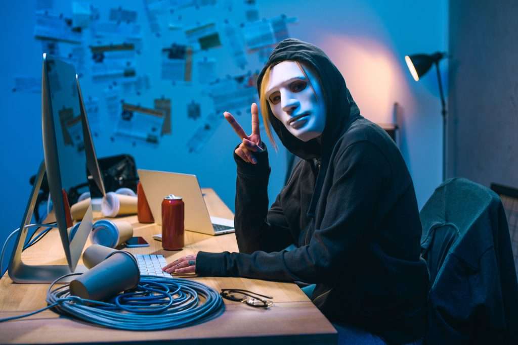 A person wearing a mask and a hoodie is seated at a desk with multiple computers and cables visible. The individual is making a peace sign with one hand while typing on a keyboard. Various items such as a can and cups are on the desk, and notes are pinned to the wall in the background.