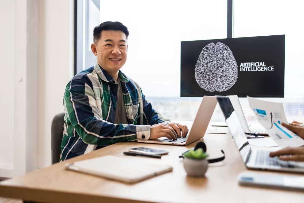 A man is sitting at a table with a laptop, smiling while working. In the background, a screen displays the words "ARTIFICIAL INTELLIGENCE" alongside a graphic. Other laptops and documents are visible on the table, suggesting a collaborative learning environment focused on artificial intelligence.