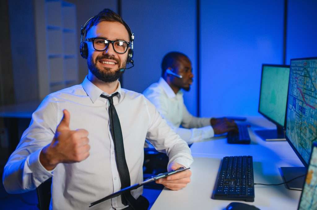 Two men are sitting at desks in a technology-focused workspace. One man, wearing glasses and a headset, is smiling and giving a thumbs-up, while the other man is focused on his computer screen. Both are engaged in an IT-related activity, possibly related to technical support or training.