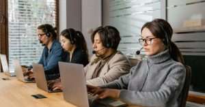 Two helpdesk technicians in an office wearing headsets and using laptops