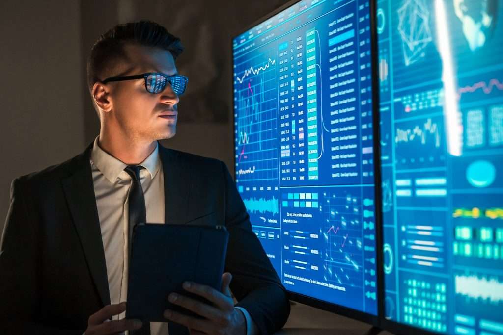A man in a suit stands in front of large screens displaying data visualizations and graphs, holding a tablet. The screens show various charts and metrics, suggesting a focus on data analysis or business intelligence.