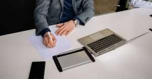 A team of professionals in an office engaging in a discussion while one writes on a laptop.