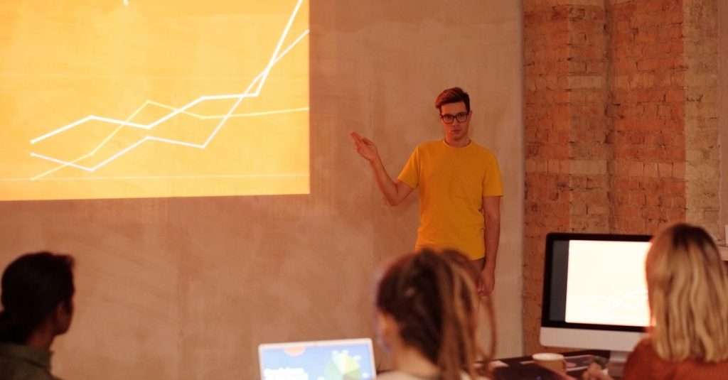 A professional presenting data on a laptop to an audience in an office setting
